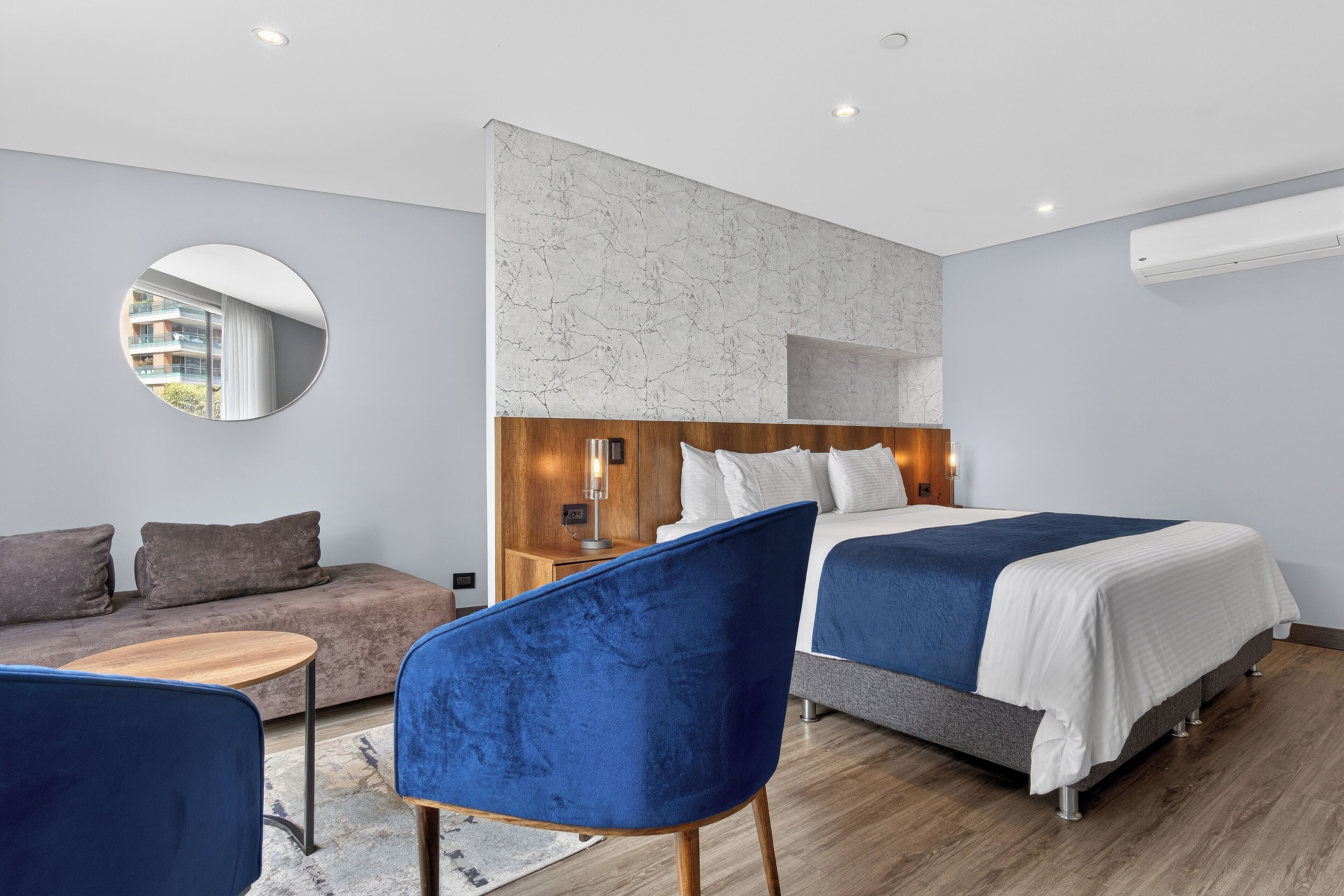 Una camera d'albergo moderna con un letto, un divano, uno specchio rotondo e una poltrona blu.