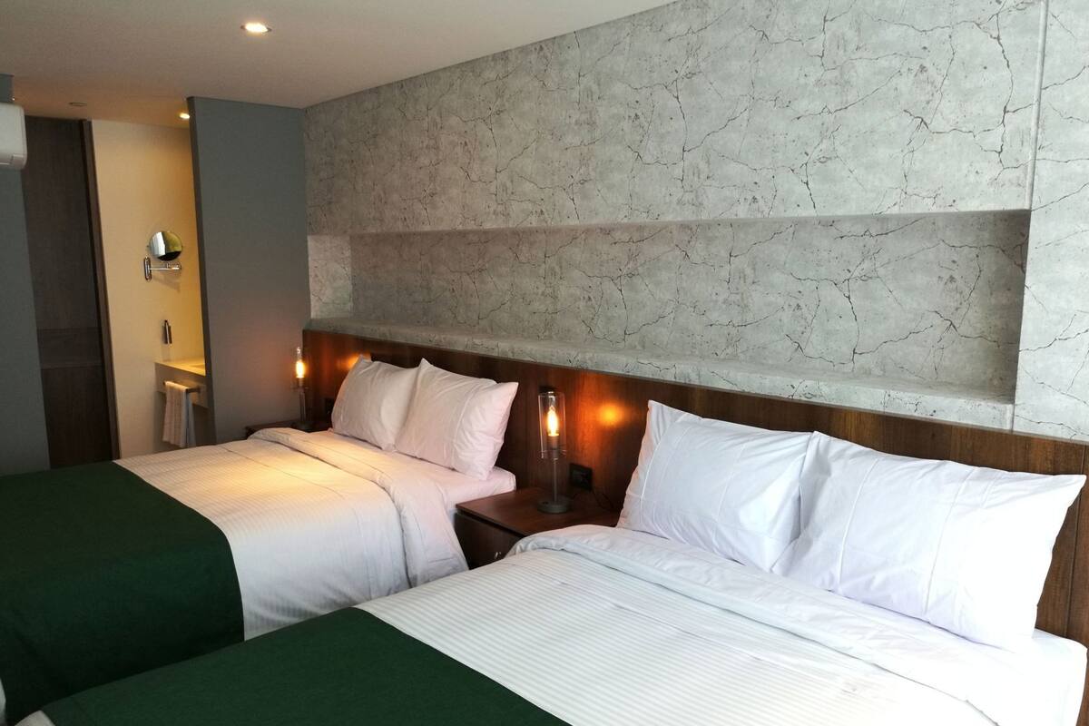 A hotel room with two beds, a marble wall, a nightstand with a lamp, and a bathroom visible in the background.
