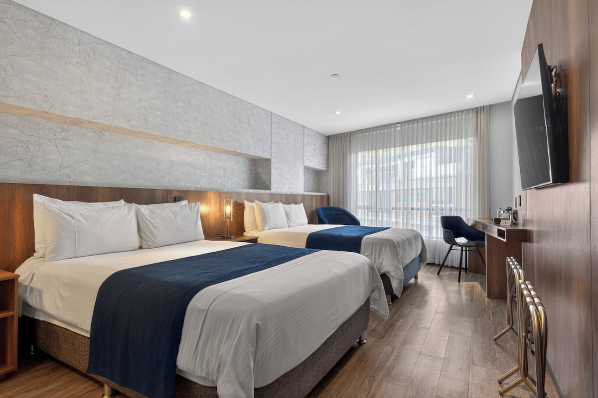 A hotel room with two beds, a desk, a chair, a TV, and a large window with curtains.