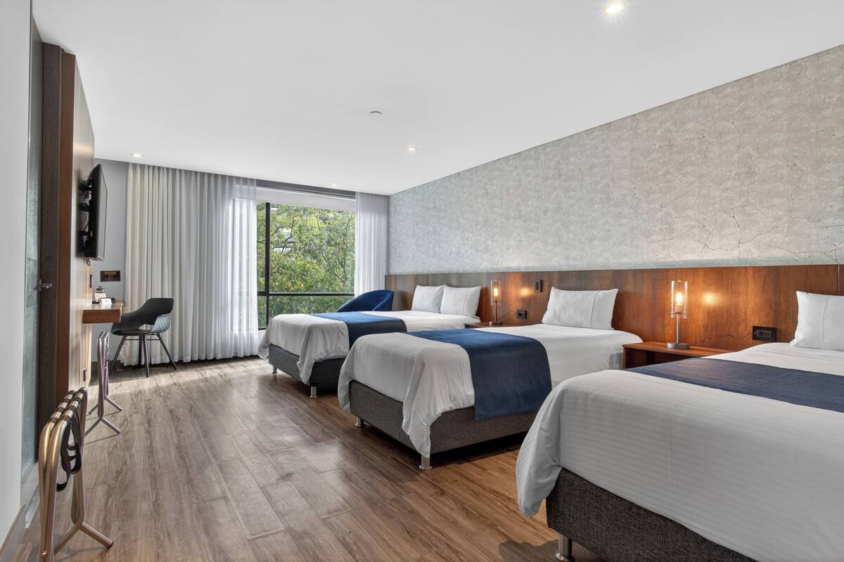 A hotel room with three beds, a desk, a chair, and a large window.