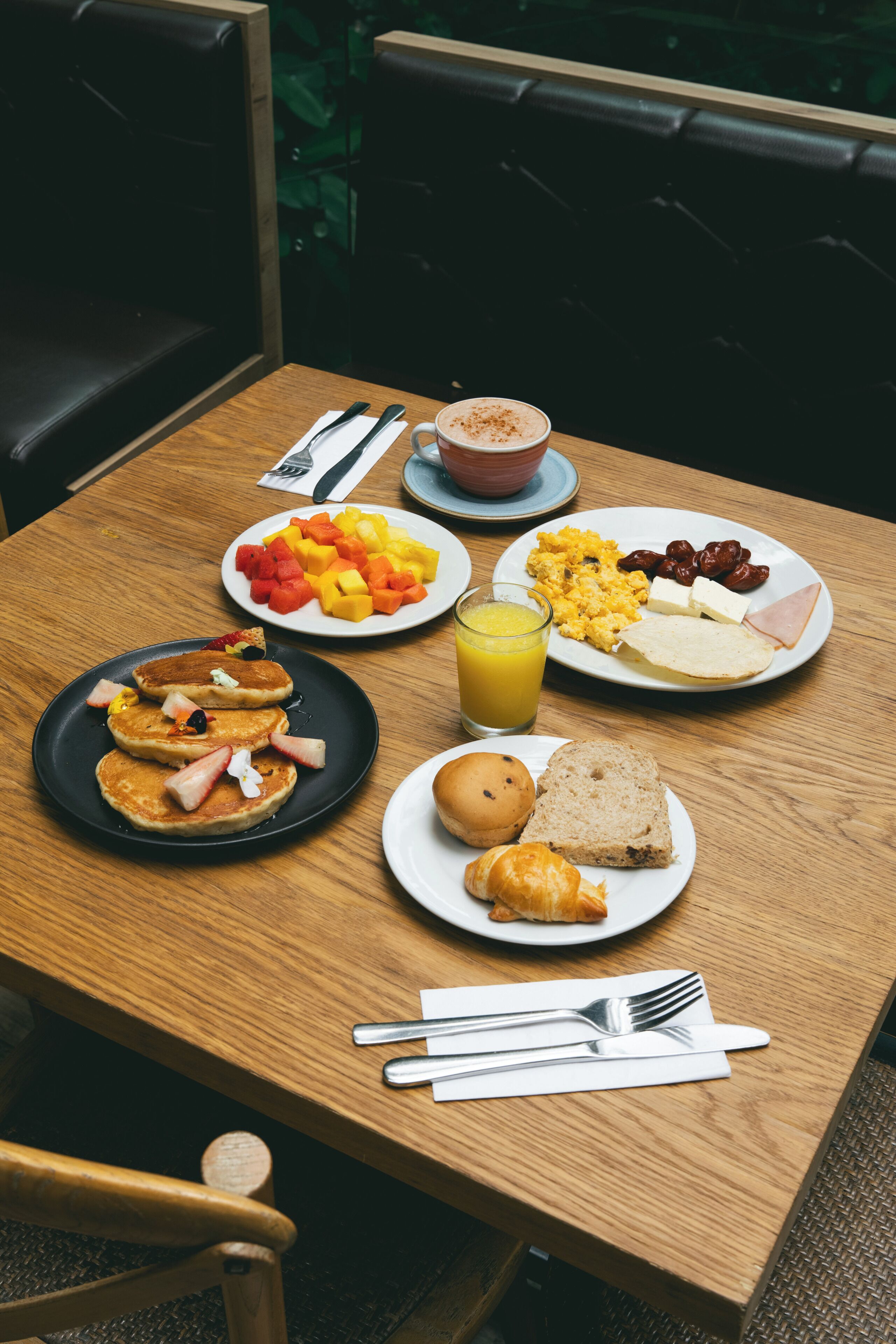 A wooden table with breakfast plates containing pancakes, eggs, fruit, and toast.