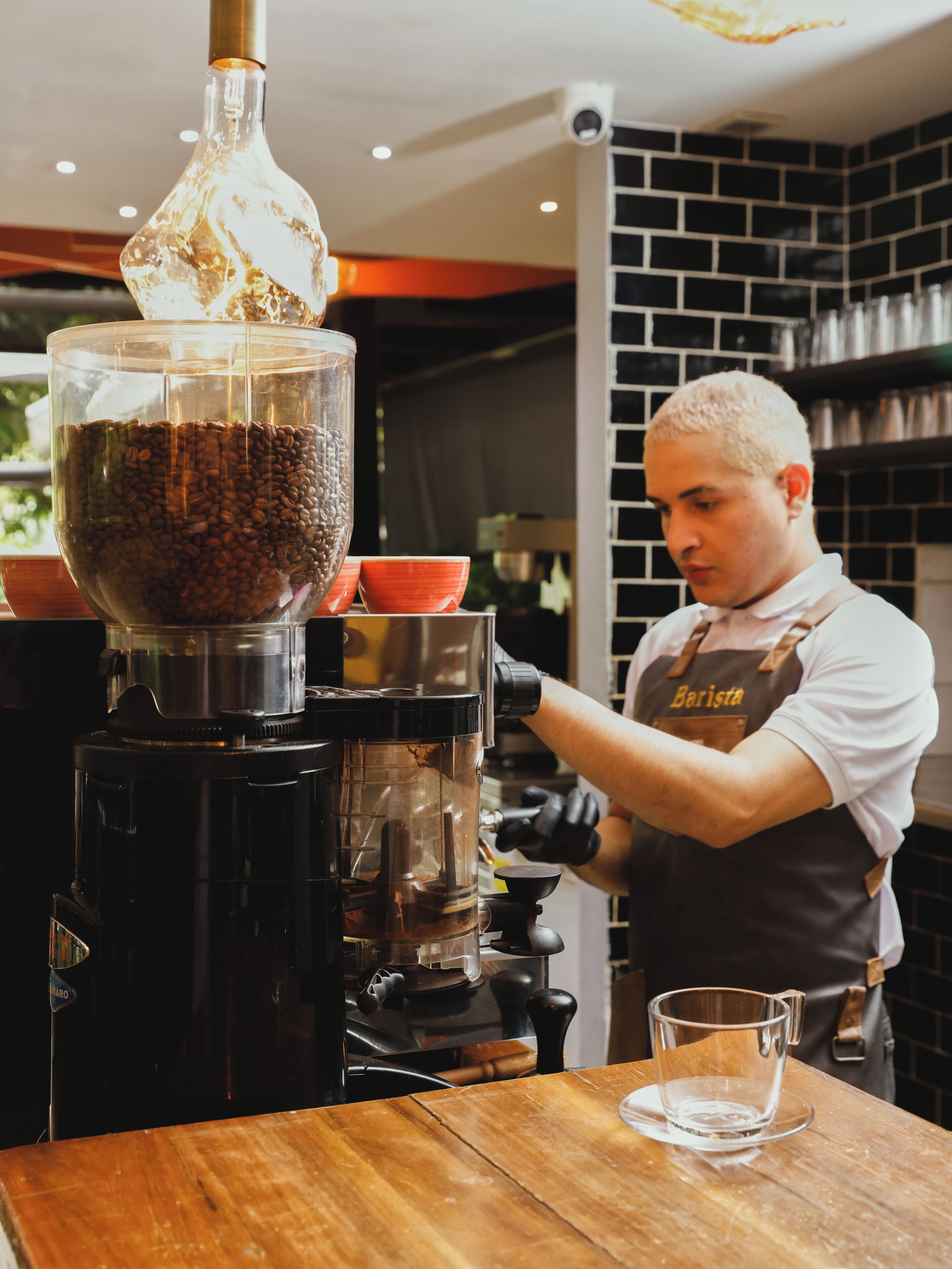 Una persona con camisa y delantal blanco operando una máquina de café con un molinillo y un recipiente de vidrio.