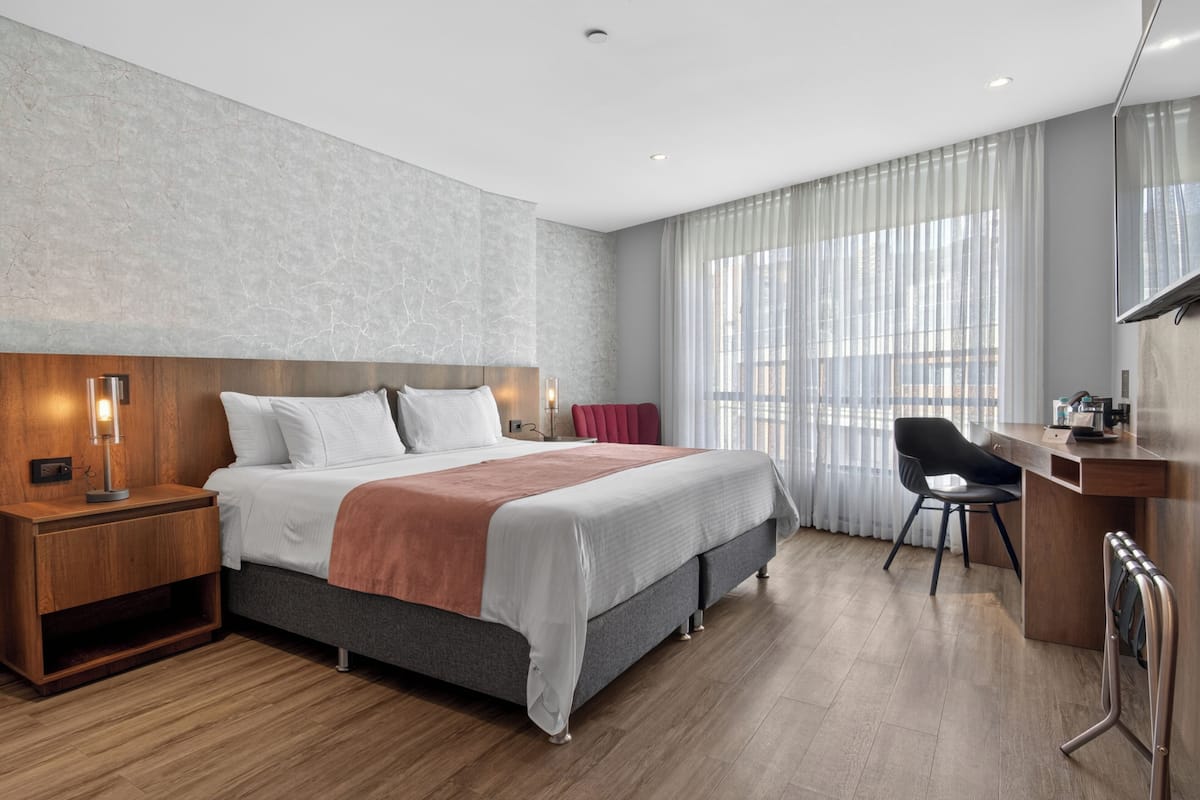 A modern hotel room with a large bed, wooden nightstand, desk, and chair.