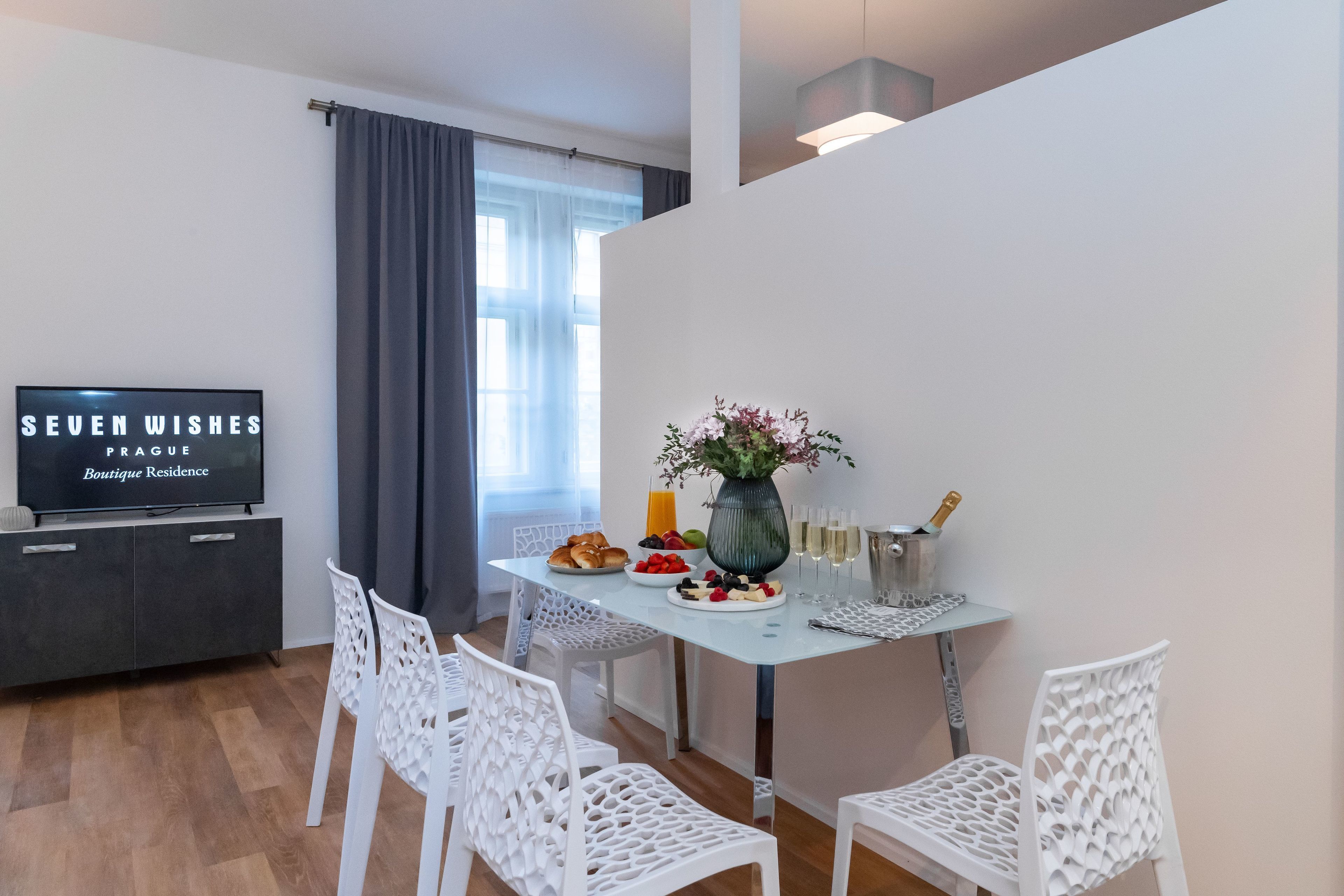 Un comedor moderno con una mesa preparada para dos personas, un florero con flores y un televisor mostrando “SEVEN WISHES PRAGUE Boutique Residence”.