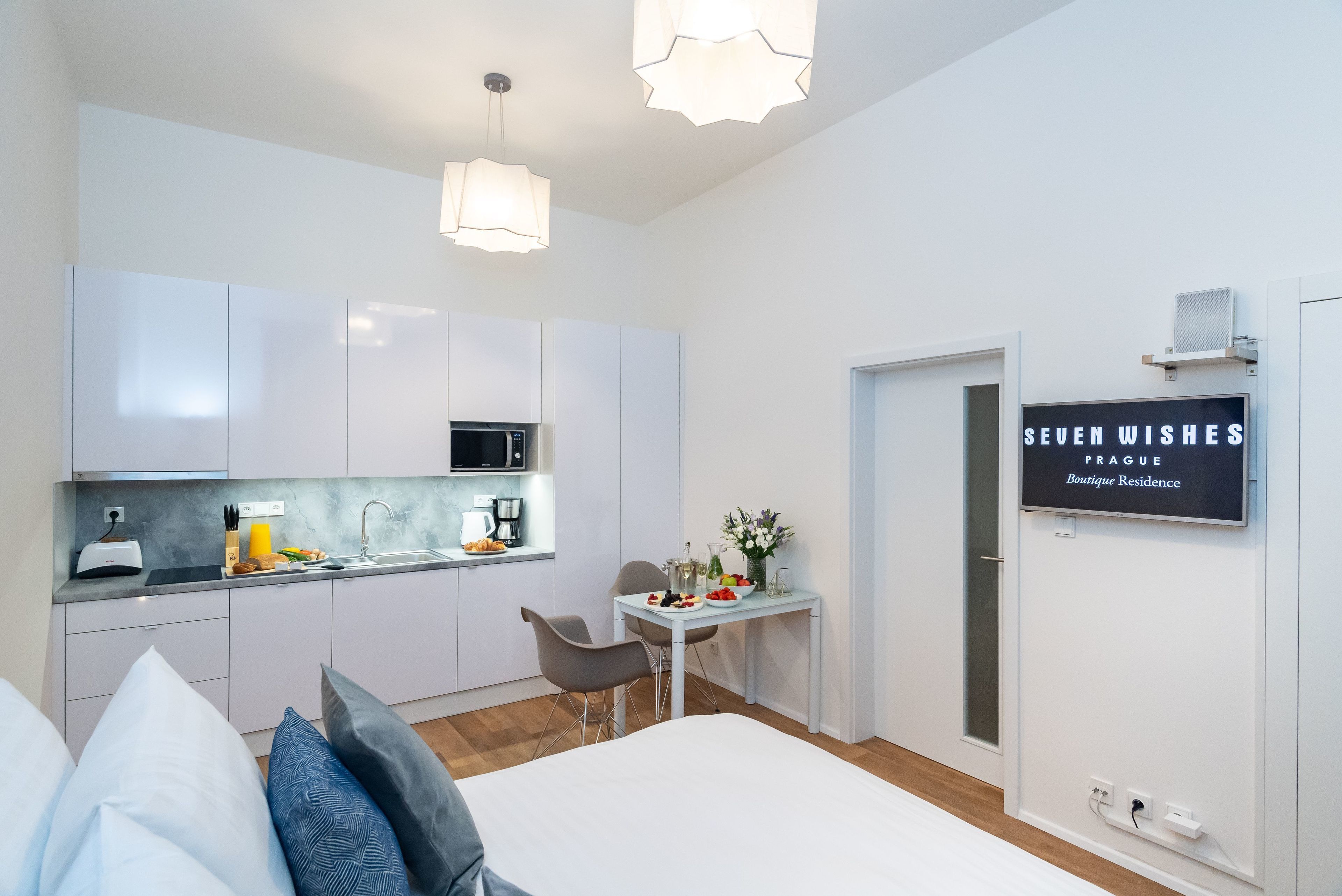 Une chambre moderne et compacte, équipée d’un lit, de tables de chevet et d’un petit bureau avec une chaise. On aperçoit une kitchenette avec un micro-ondes, un évier et des placards. Une pancarte sur le mur indique : « SEVEN WISHES PRAGUE, résidence de luxe ».