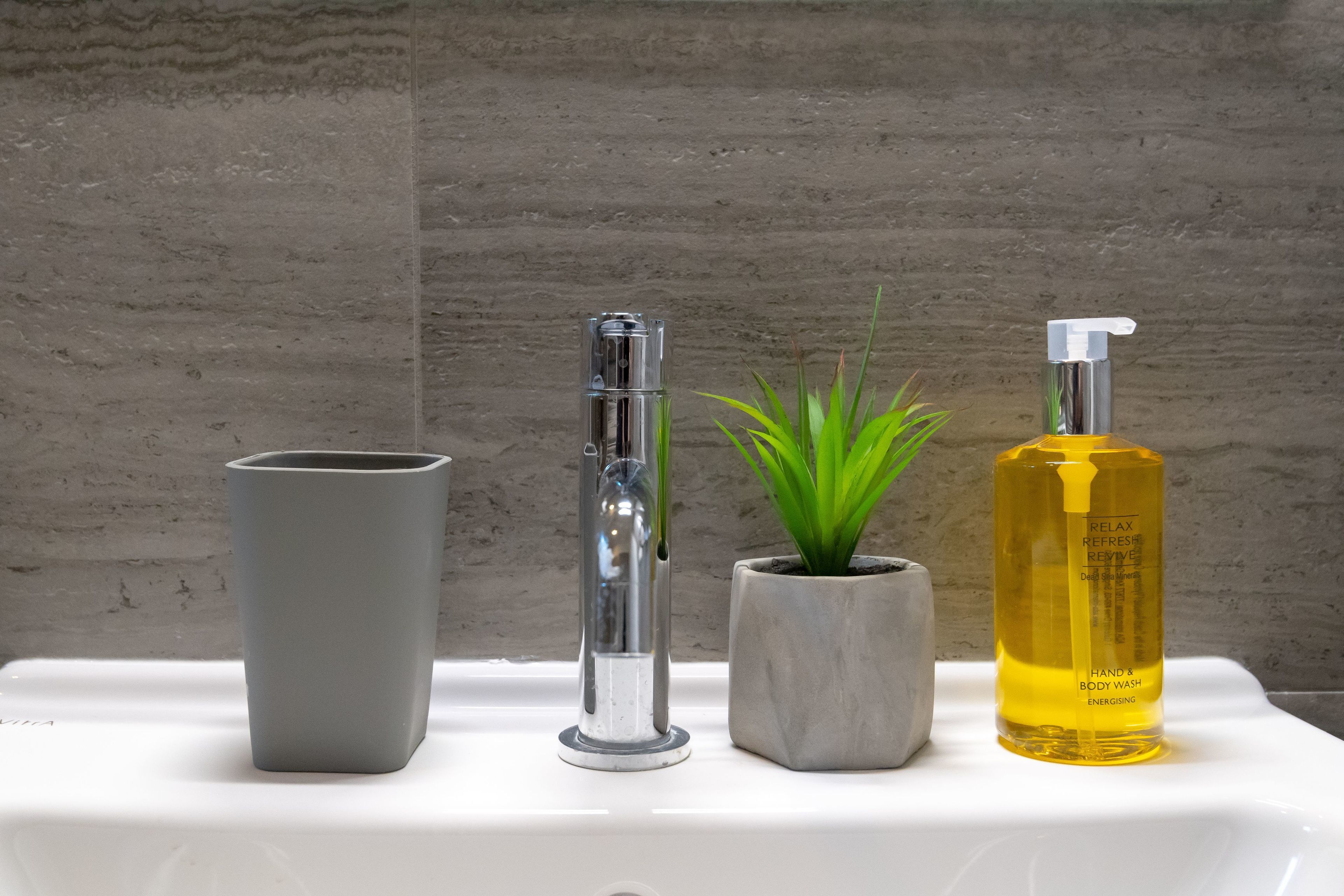 Een wastafel met een grijze vaas, een hoge chromen kraan, een potplant en een gele fles hand- en douchezeep.