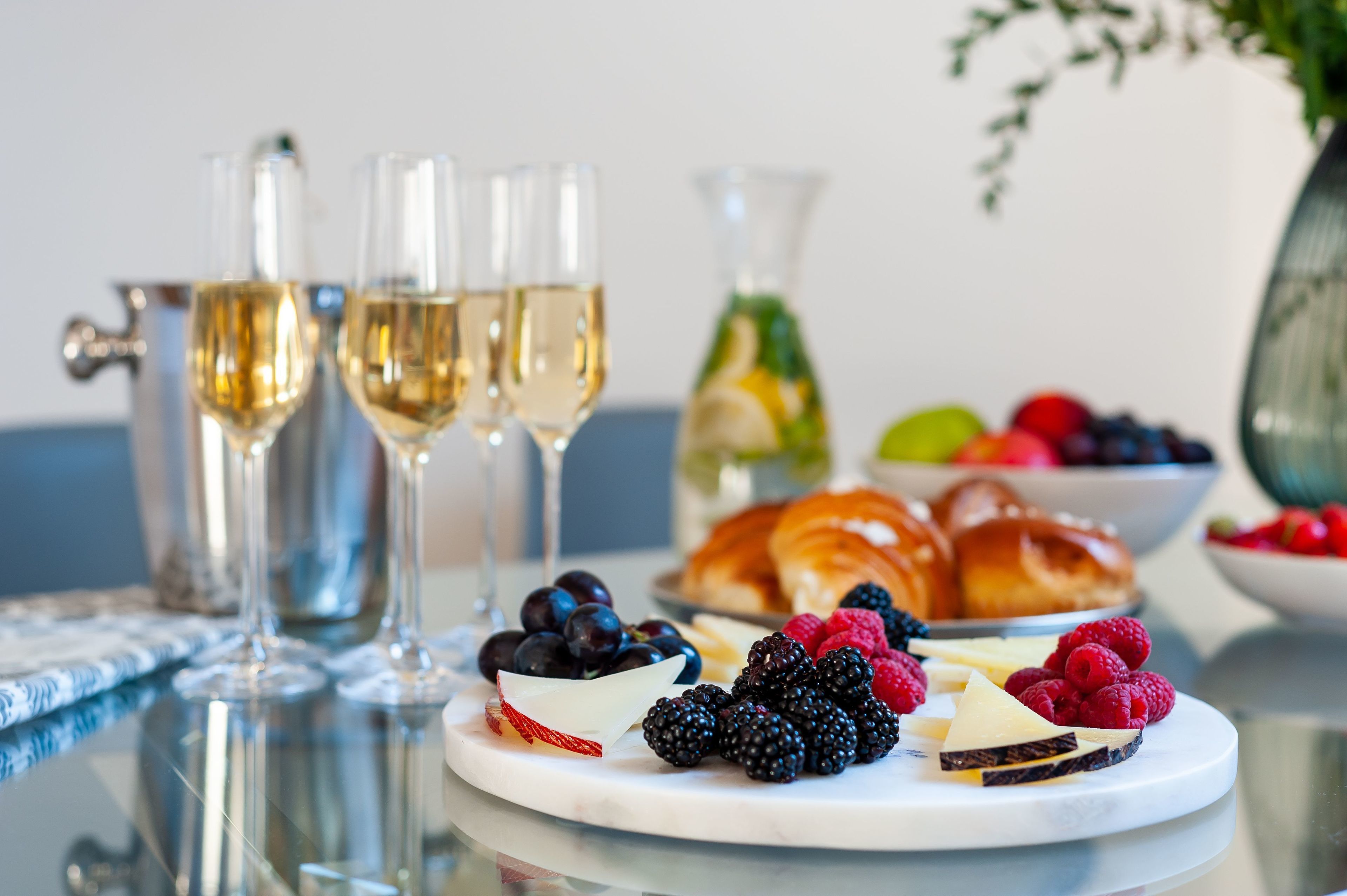 Una mesa con un plato de frutas variadas, una jarra y dos copas de vino blanco.