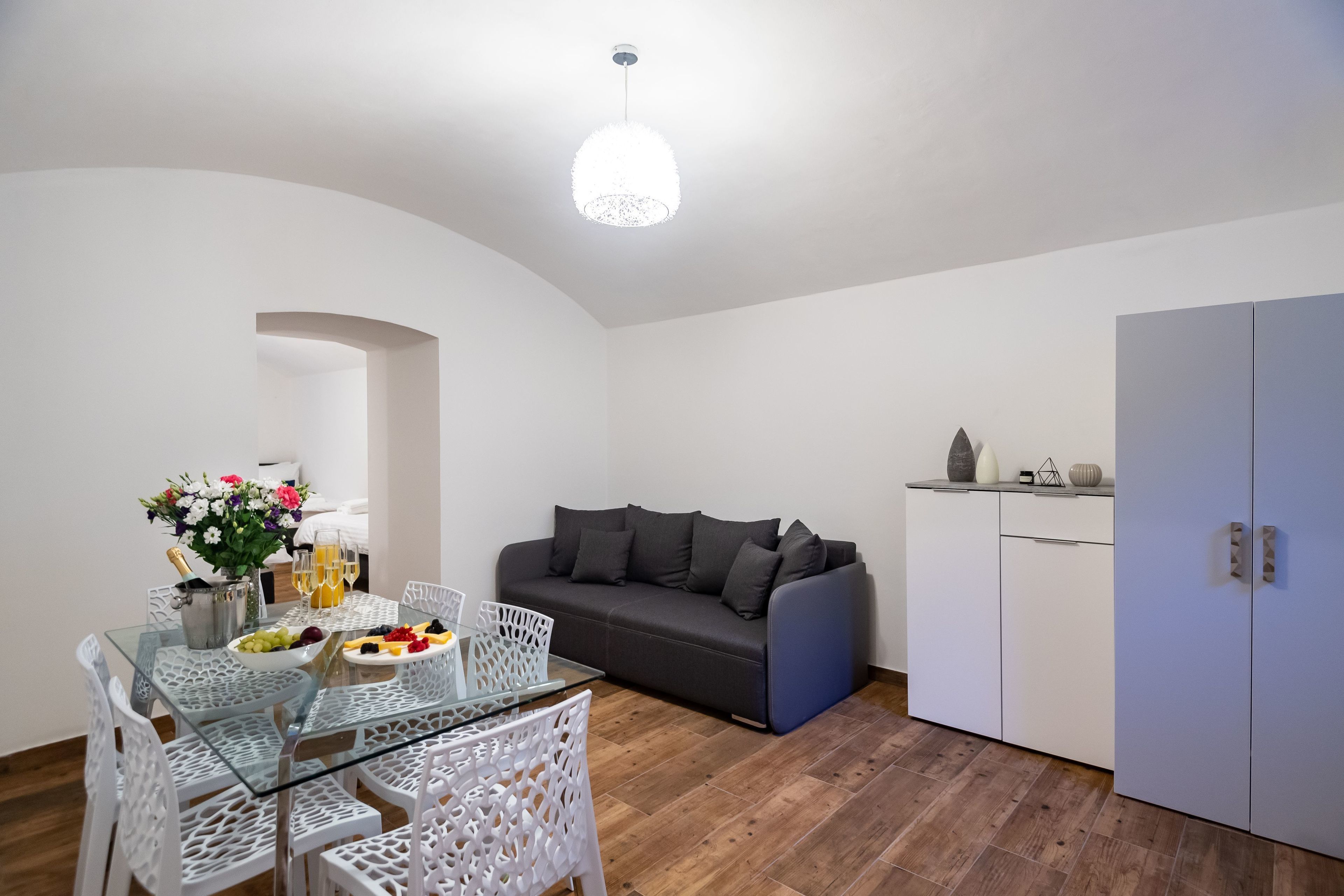 A modern living room with a glass dining table, white chairs, a grey sofa, and a cabinet.