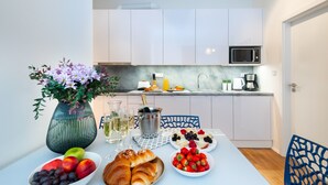 Een moderne keuken met witte kastjes, een magnetron en een koffiezetapparaat. Er staat een eettafel met fruit, gebak en een bloevas.