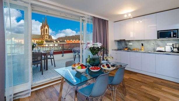 Een moderne keuken met witte kastjes en een magnetron, een eetgedeelte met een glazen tafel en stoelen, en een uitzicht op een kerk door grote ramen.