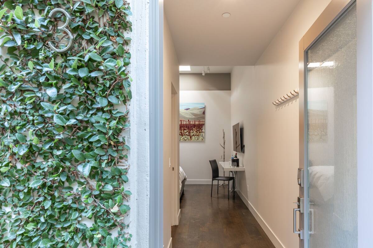 A hallway with a wall covered in green foliage, a desk with a chair, and a bed in the background.