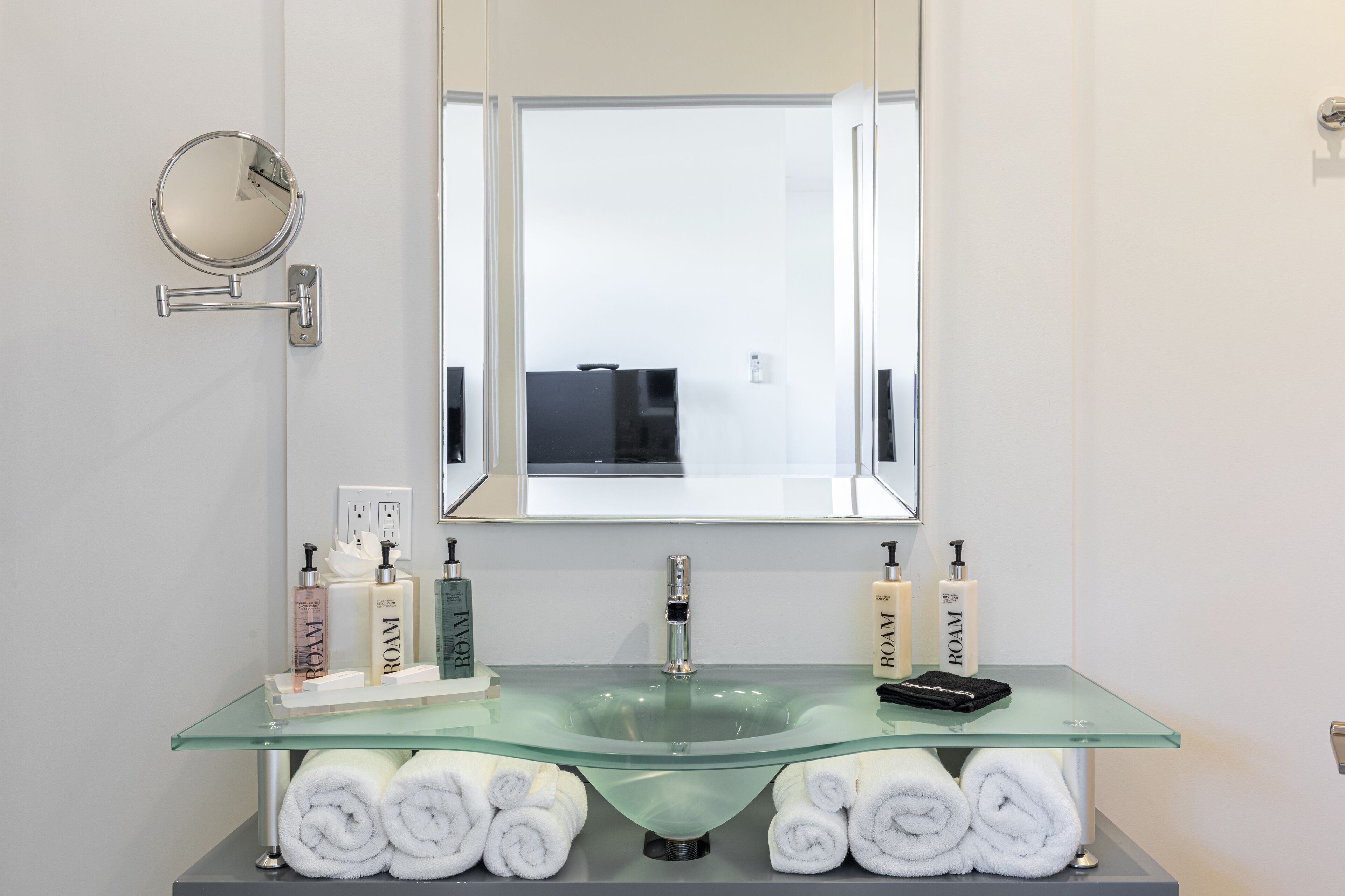 Un baño con un lavabo de vidrio, dos botellas de ROAM, un espejo redondo y toallas.