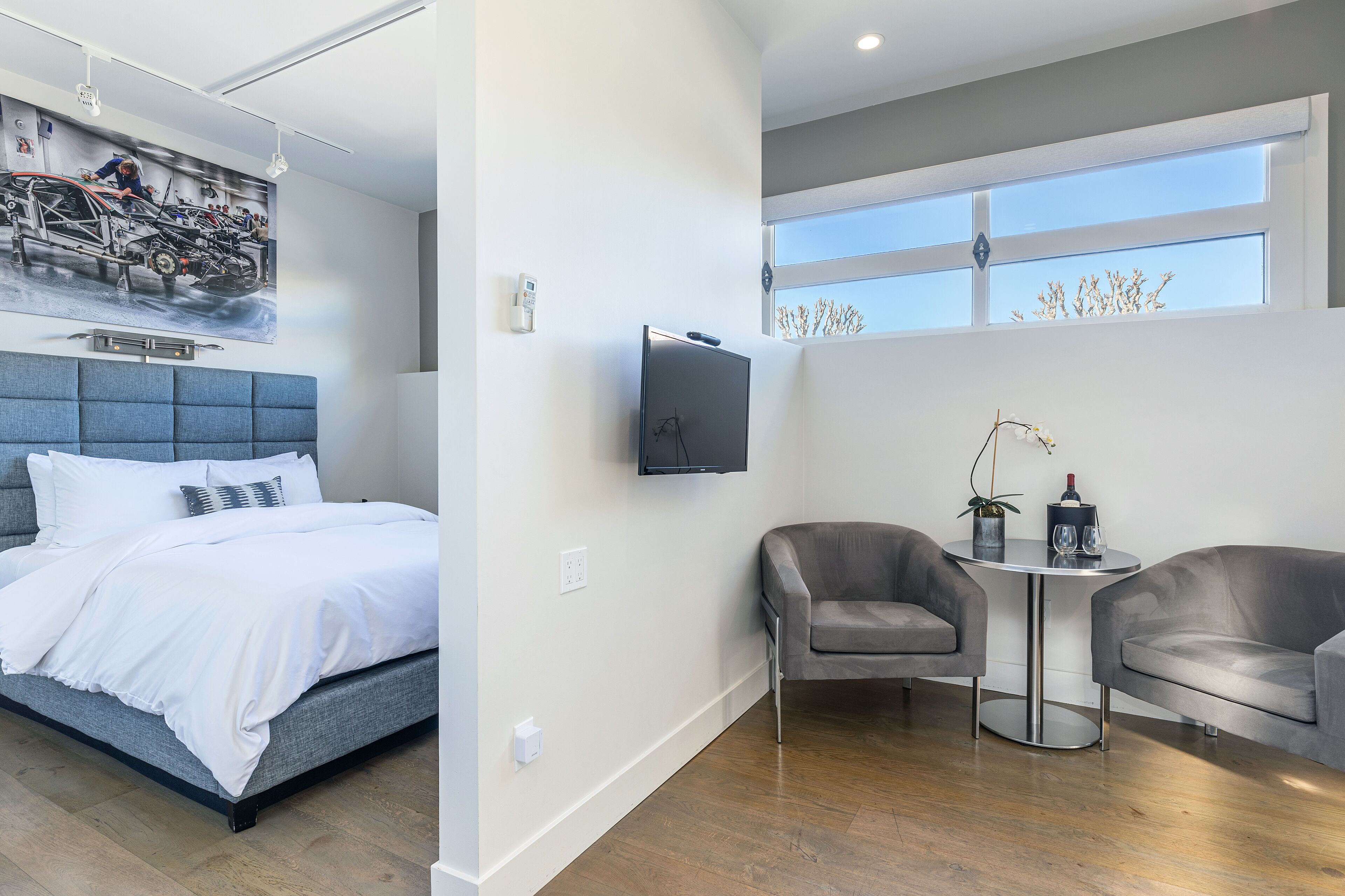 A modern hotel room with a bed, a TV, a seating area with two chairs, and a large window.