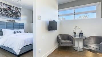 A modern hotel room with a bed, a TV, a seating area with two chairs, and a large window.