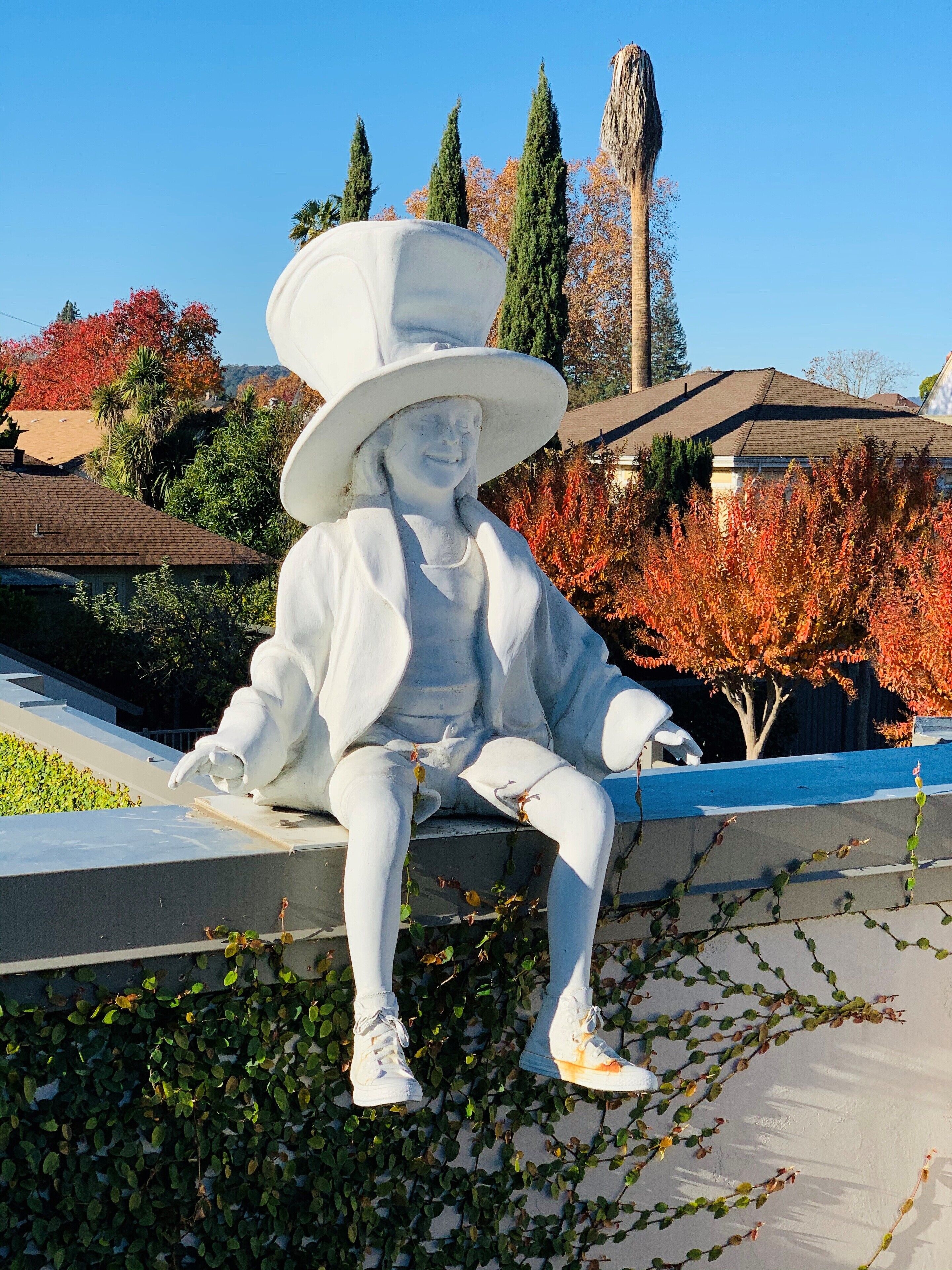 Una estatua de una persona con un sombrero grande sentada en un saliente, con un jardín y árboles de fondo.