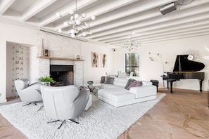 Una sala de estar moderna con un sofá blanco, una mesa de centro de vidrio, una chimenea, un piano de cola y un ventanal amplio.