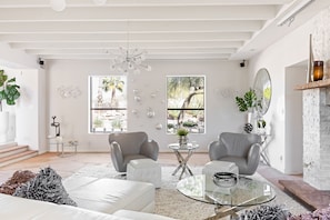 Una sala de estar moderna con dos sillones grises, un sofá blanco, una mesa de centro de vidrio y una chimenea. Hay dos ventanas con vistas a la vegetación, y un techo con una lámpara de techo original.