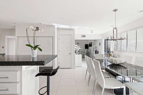A modern kitchen with white cabinetry, a black countertop, and a dining area with white chairs and a glass table.