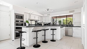 Una cocina moderna con armarios blancos, una isla central con taburetes de bar, electrodomésticos de acero inoxidable y un amplio ventanal con vistas a zonas verdes.