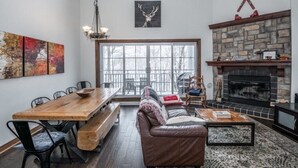 A spacious living room with a large wooden dining table, a leather sofa, a fireplace, and a large window with a view of trees. - Cozy condo - Walk to village/mountain - Monthly rates (Mont-Tremblant)