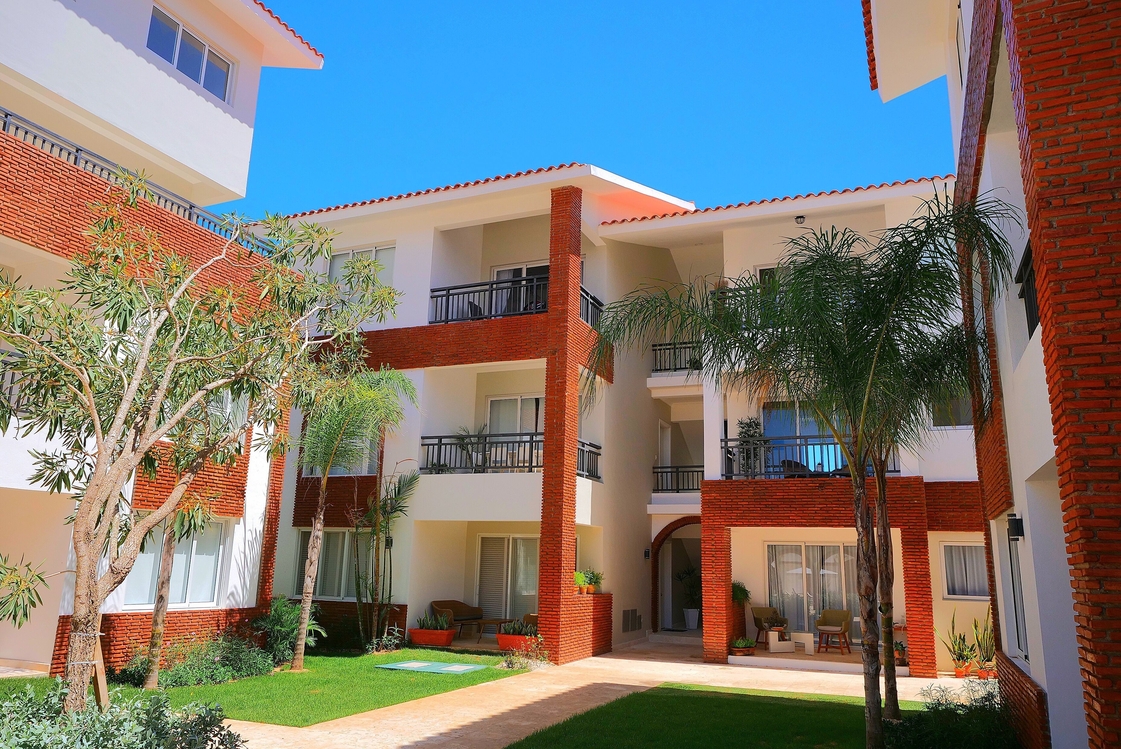 A multi-story residential building with balconies, a central courtyard with greenery, and a clear blue sky.