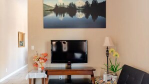 Een woonkamer met een flatscreen televisie op een houten standaard, een bank, een kleine tafel met een bloevas en een schilderij van een meer aan de muur.