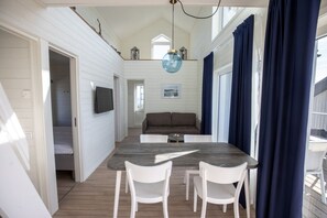 A compact room with a wooden floor, a dining table with two chairs, a sofa, a flat-screen TV mounted on the wall, and a ceiling light fixture.
