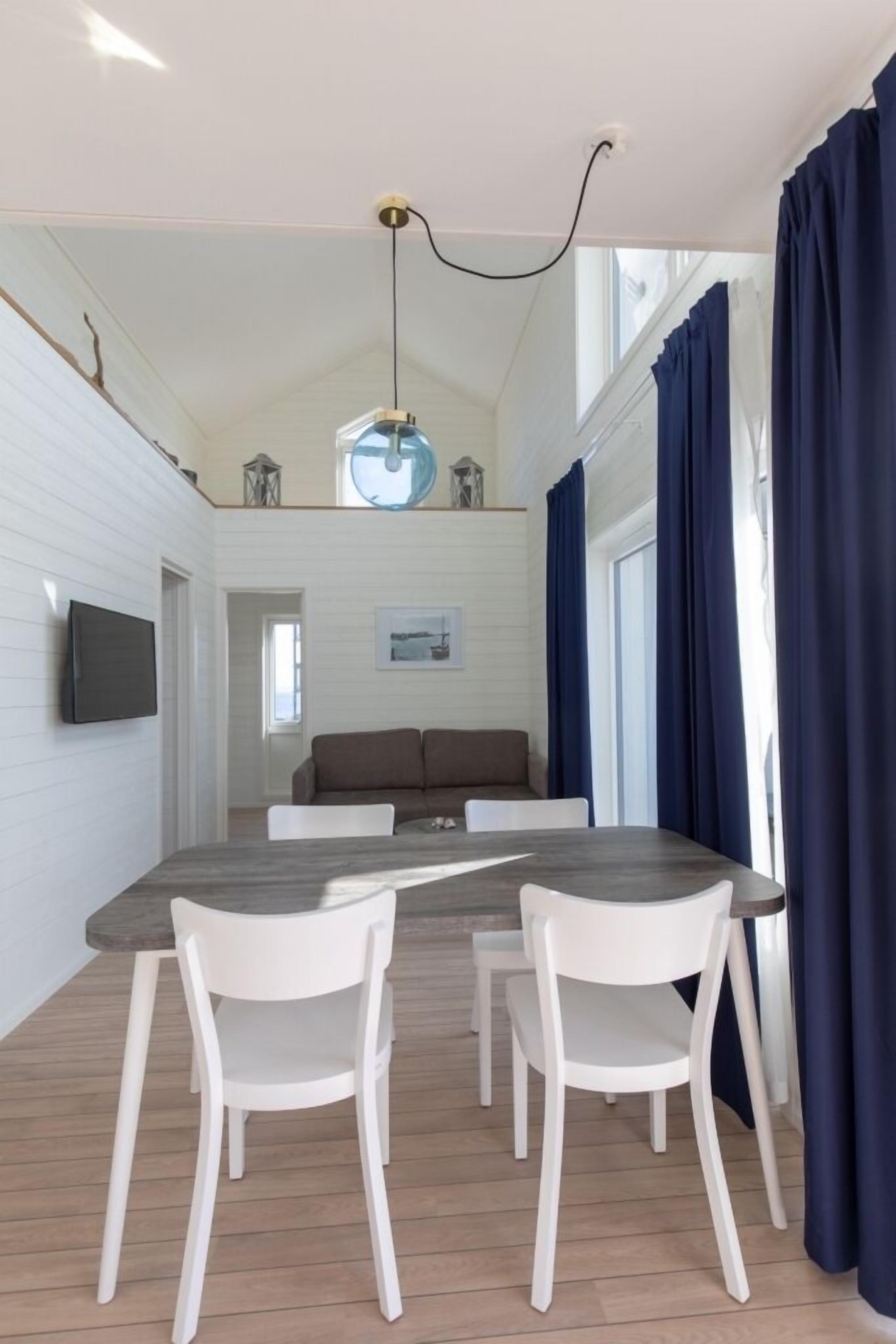 A dining area with a wooden table, white chairs, a ceiling light, and a view of a balcony with blue curtains.
