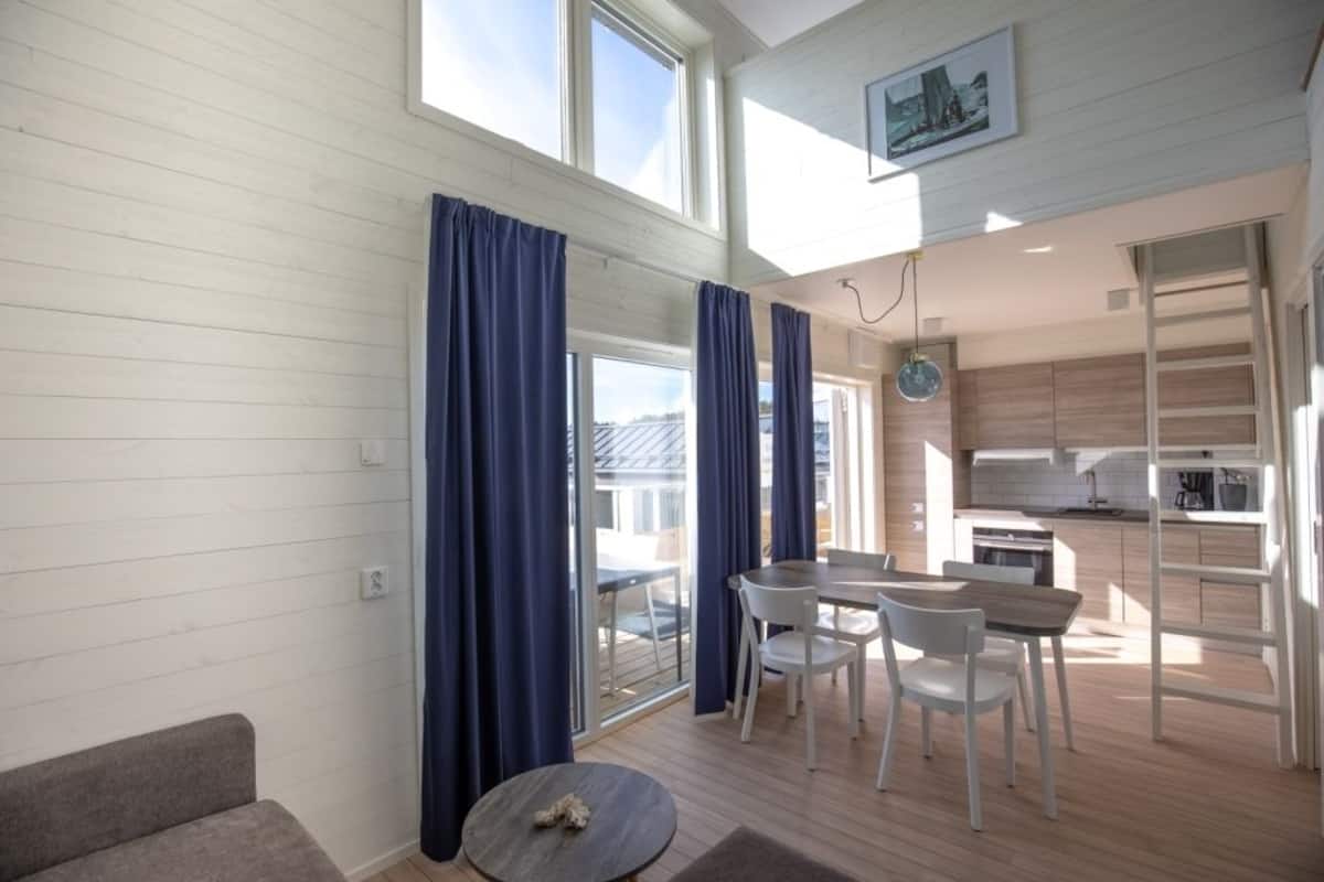 A modern living room with a sofa, a dining table with chairs, and a small round table with a decorative item. There is a kitchen area with wooden cabinets and a window overlooking a deck.