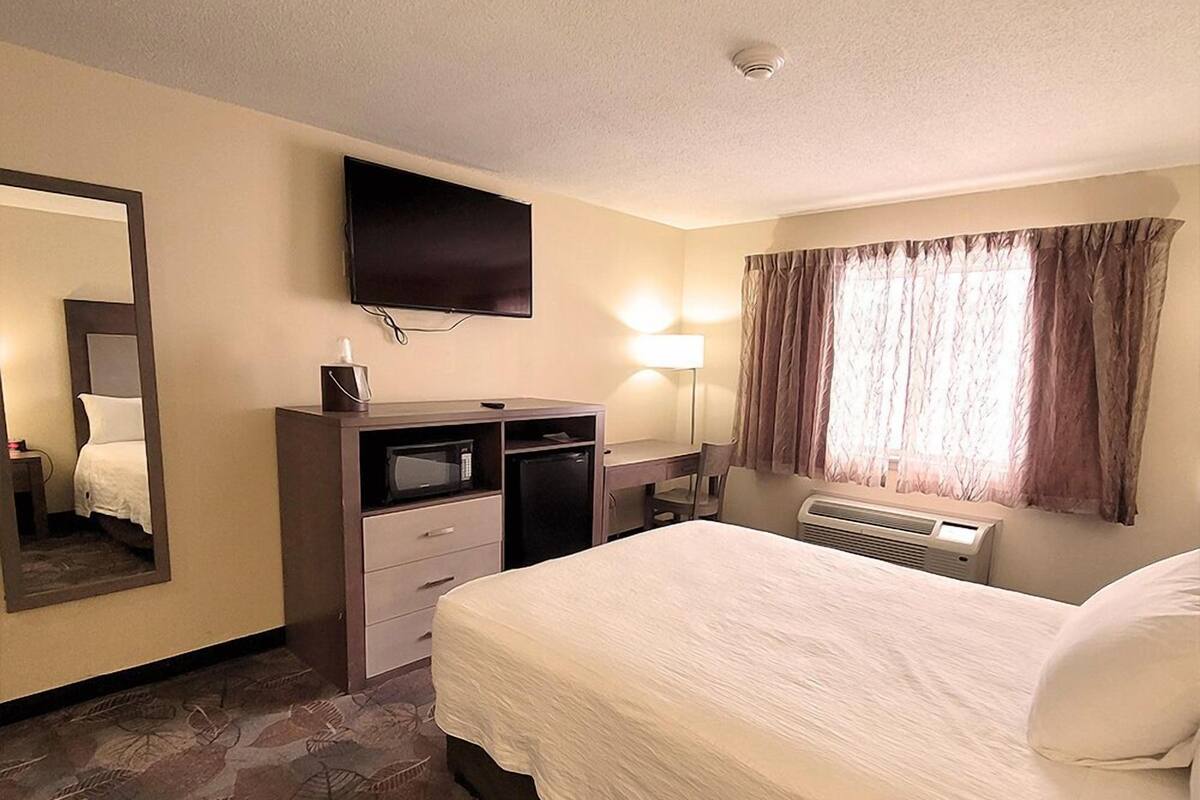 A hotel room with a bed, a desk with a computer, a television mounted on the wall, a window with curtains, and a ceiling vent.