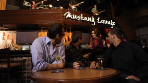 Zona de bar con un letrero de neón que dice "Mustang Lounge", un escenario con músicos y clientes sentados en mesas con bebidas.