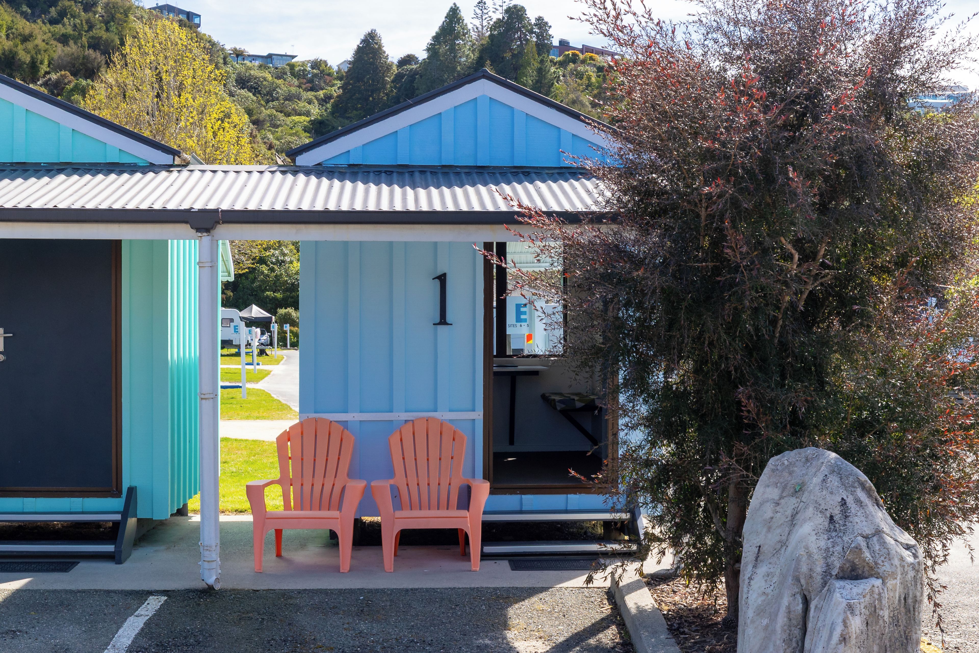A small, single-story building with a blue roof and a number '1' sign, indicating a lodging unit. There are two chairs outside the entrance, and a large rock is visible to the right.