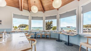 A dining area with wooden tables and chairs, large windows with ocean views, and hanging paper lanterns. - Kaiteriteri Reserve Cabins (Kaiteriteri)
