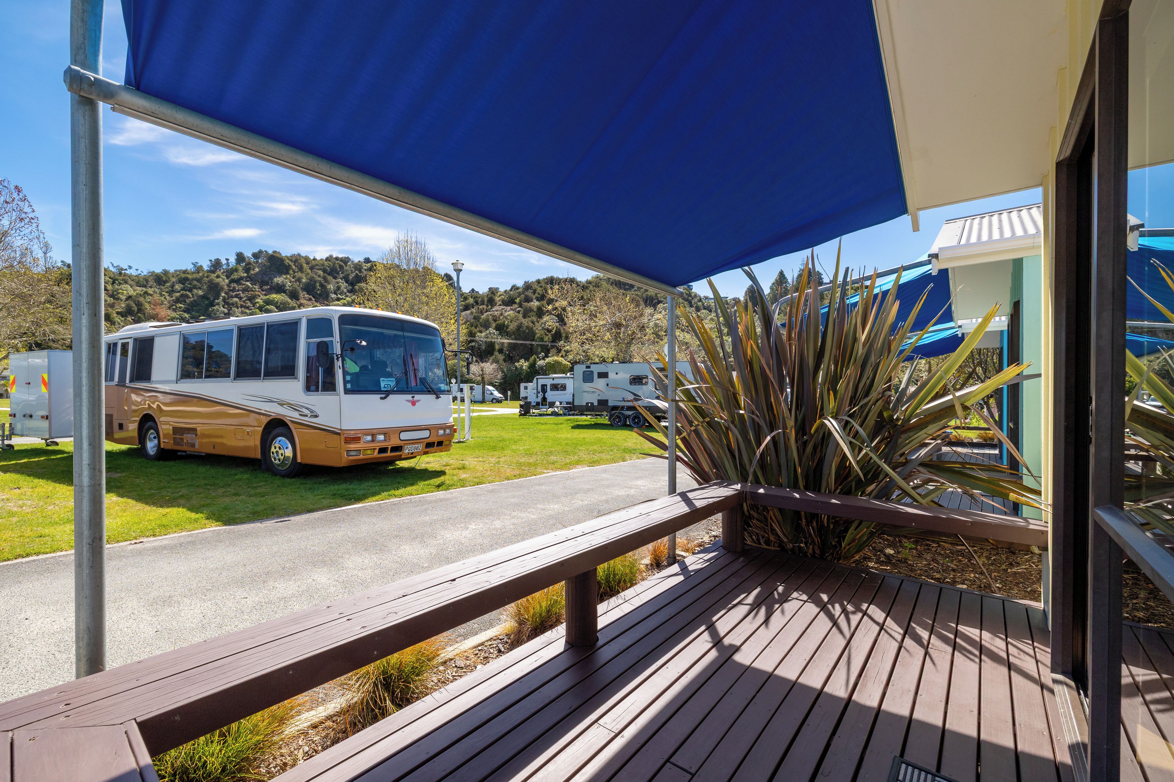 A covered outdoor area with a bus and RVs parked, a wooden deck, and a view of a hilly landscape.
