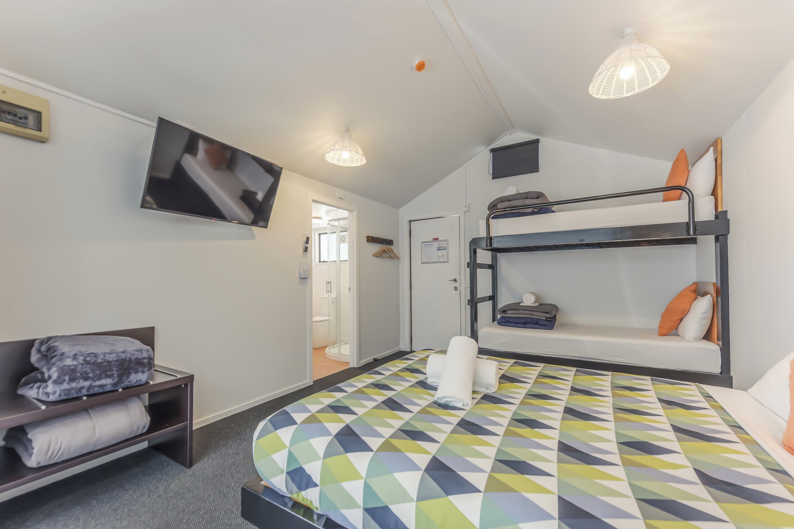 A hotel room with a bunk bed, a flat-screen TV mounted on the wall, a bedside table with towels, and a patterned bedspread.