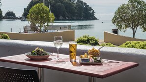 A table with a bowl of salad, a glass of white wine, a can of beer, and a plate of fries, overlooking a body of water with boats and trees. - Kaiteriteri Reserve Cabins (Kaiteriteri)