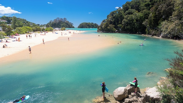A beach with clear turquoise water, people sunbathing, and a few surfers. - Kaiteriteri Reserve Cabins (Kaiteriteri)