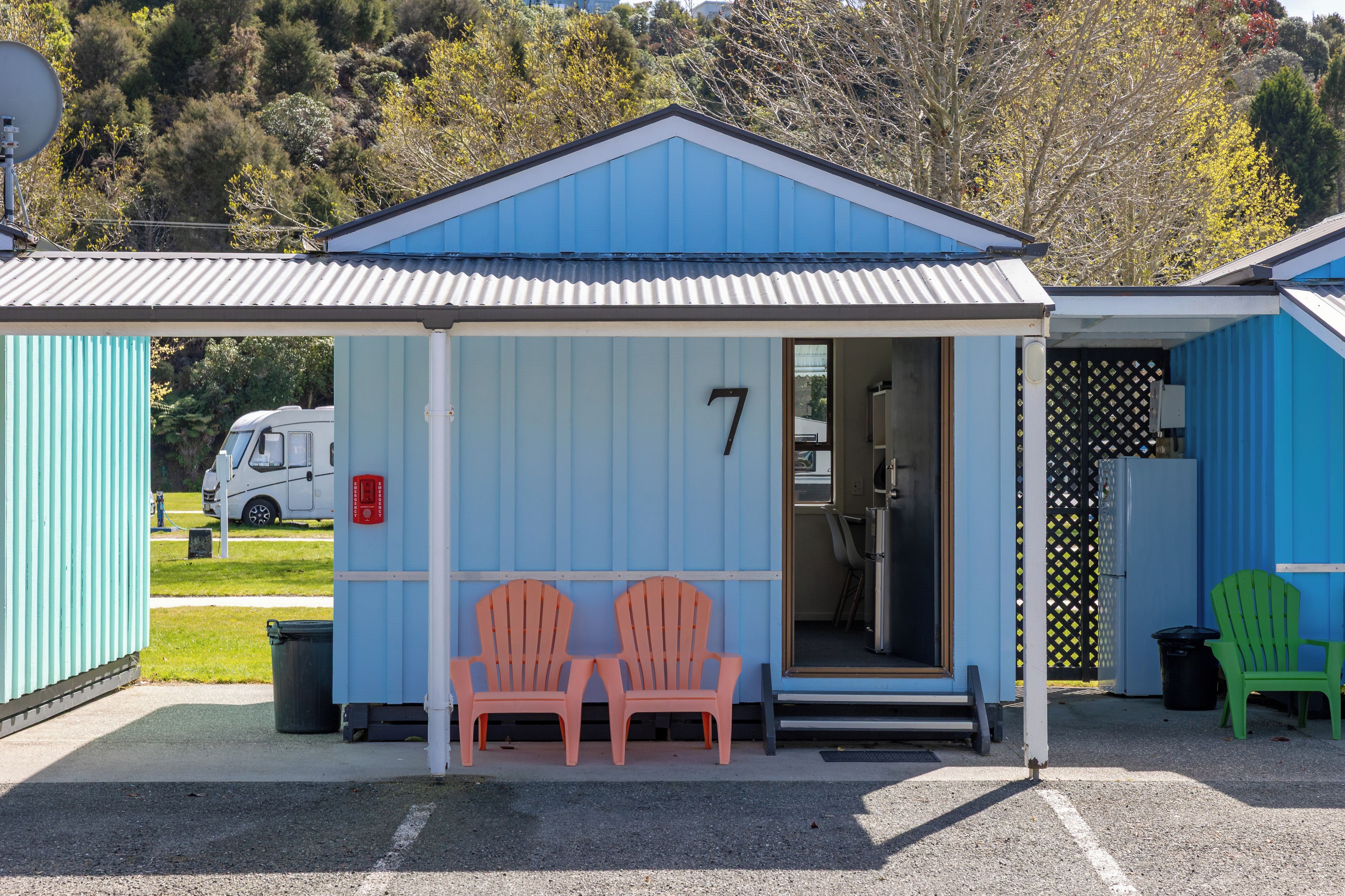 A small, single-story building with a blue exterior and a white roof. It has a number '7' on the front wall, indicating its unit number. There are two chairs outside the entrance, and a small door leading inside.