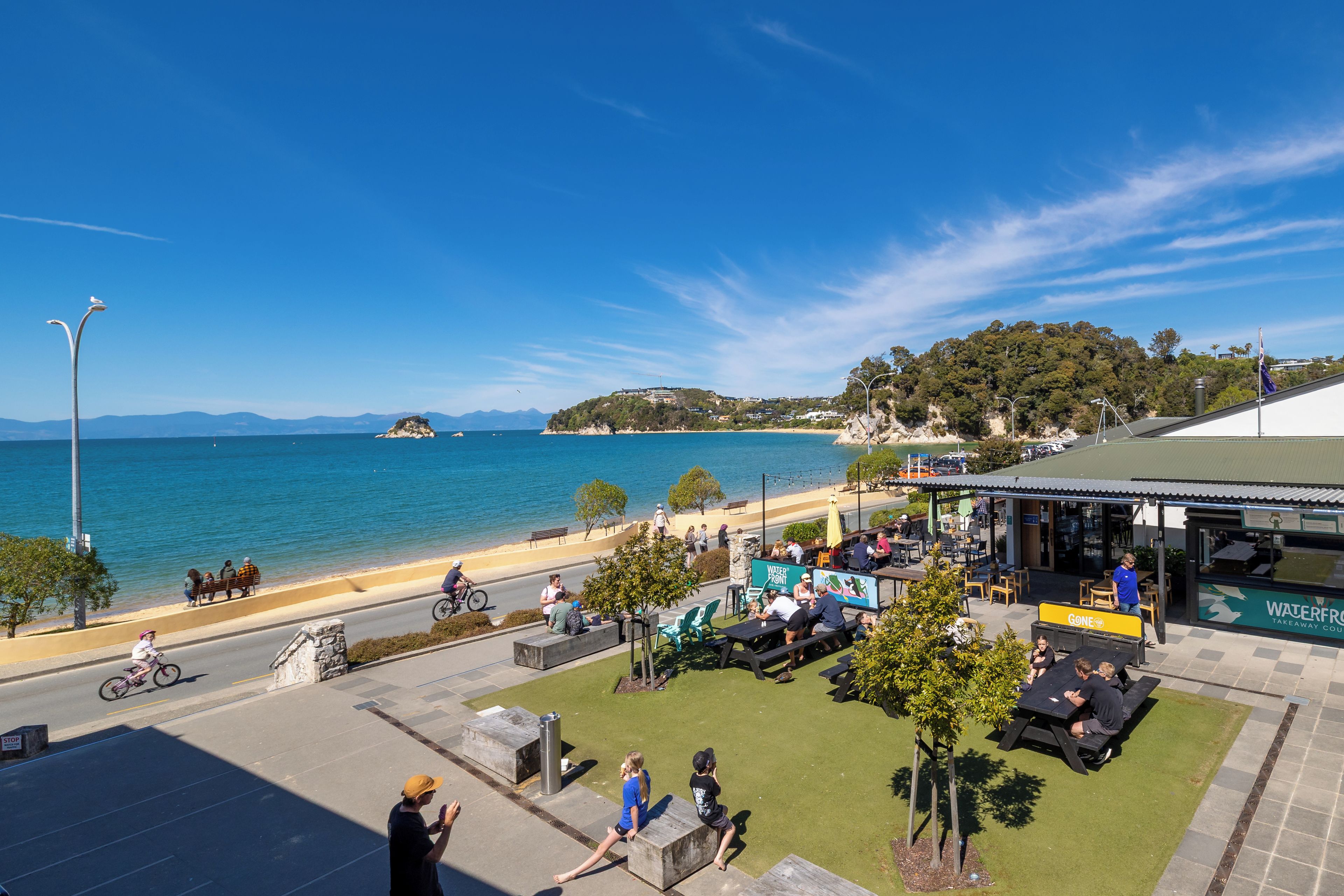 Una zona costera amb vista al mar, un cafè amb terrassa i un edifici amb un rètol que diu 'WATERFRONT TAKEAWAY CO'. 