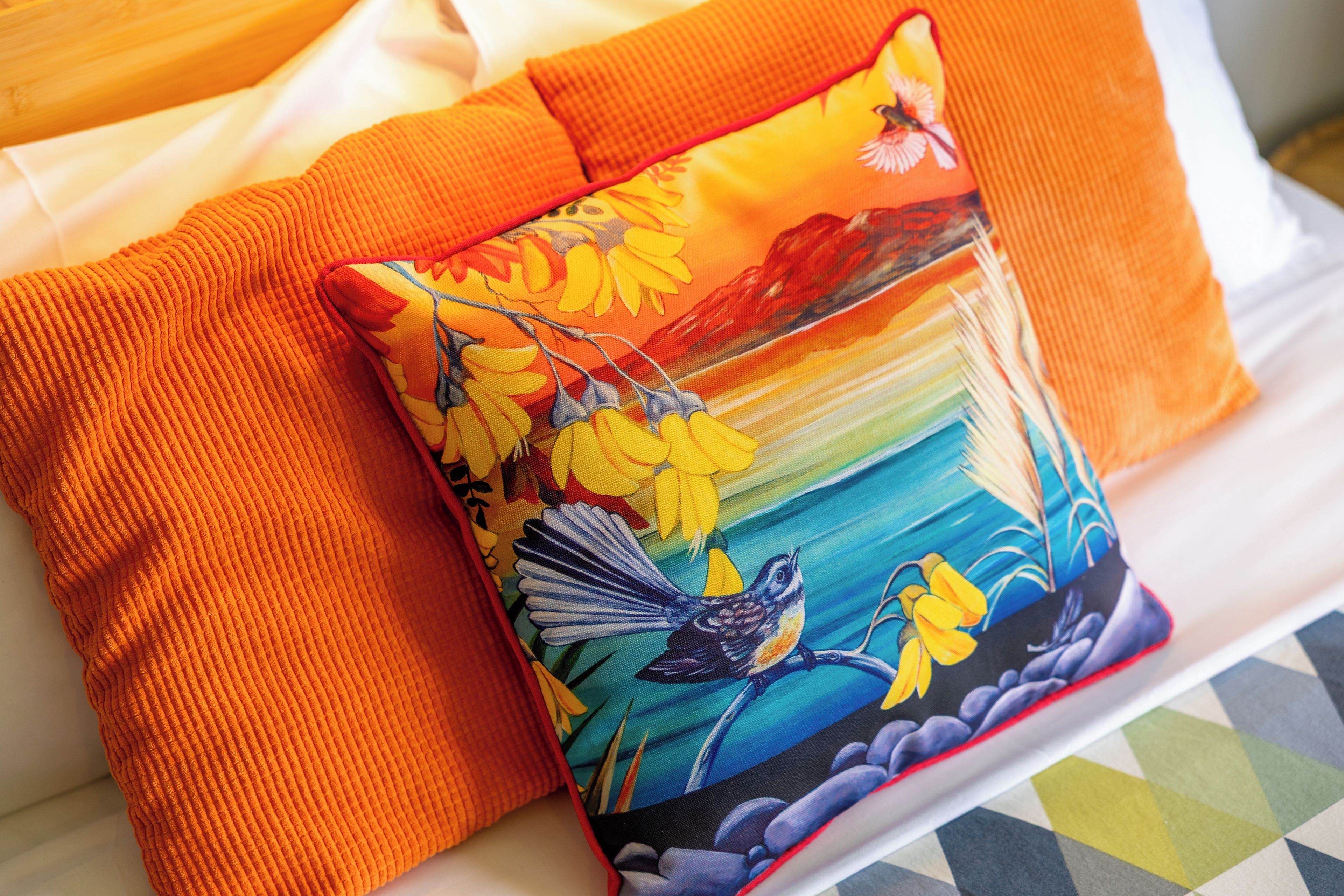 Una cama con un almohadón de colores que muestra un diseño floral y de pájaros, y una manta naranja con textura.