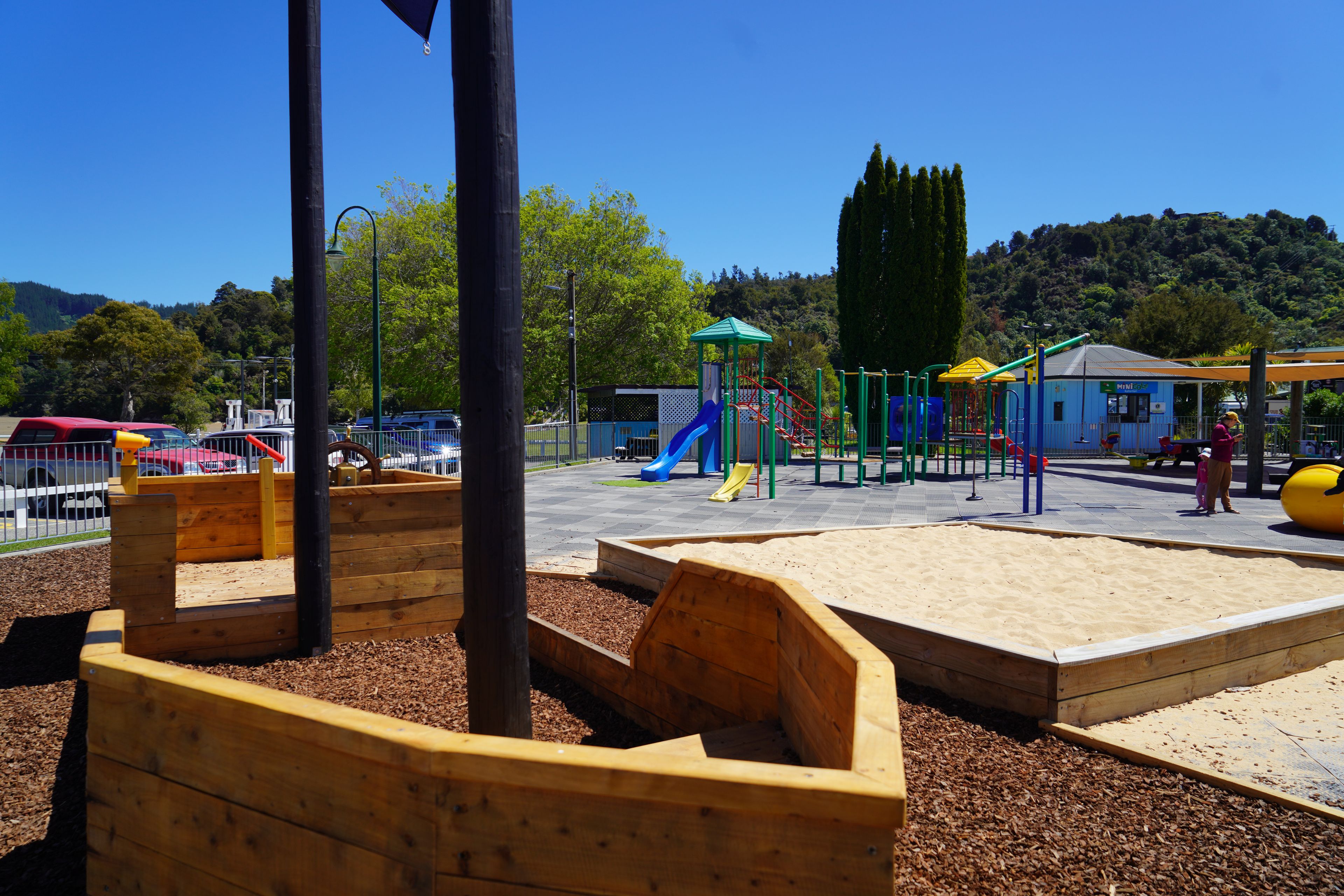 Un parque infantil con diversas estructuras de juego, incluyendo toboganes y juegos de escalada, rodeado de zonas de juego de madera y una sección de arena.