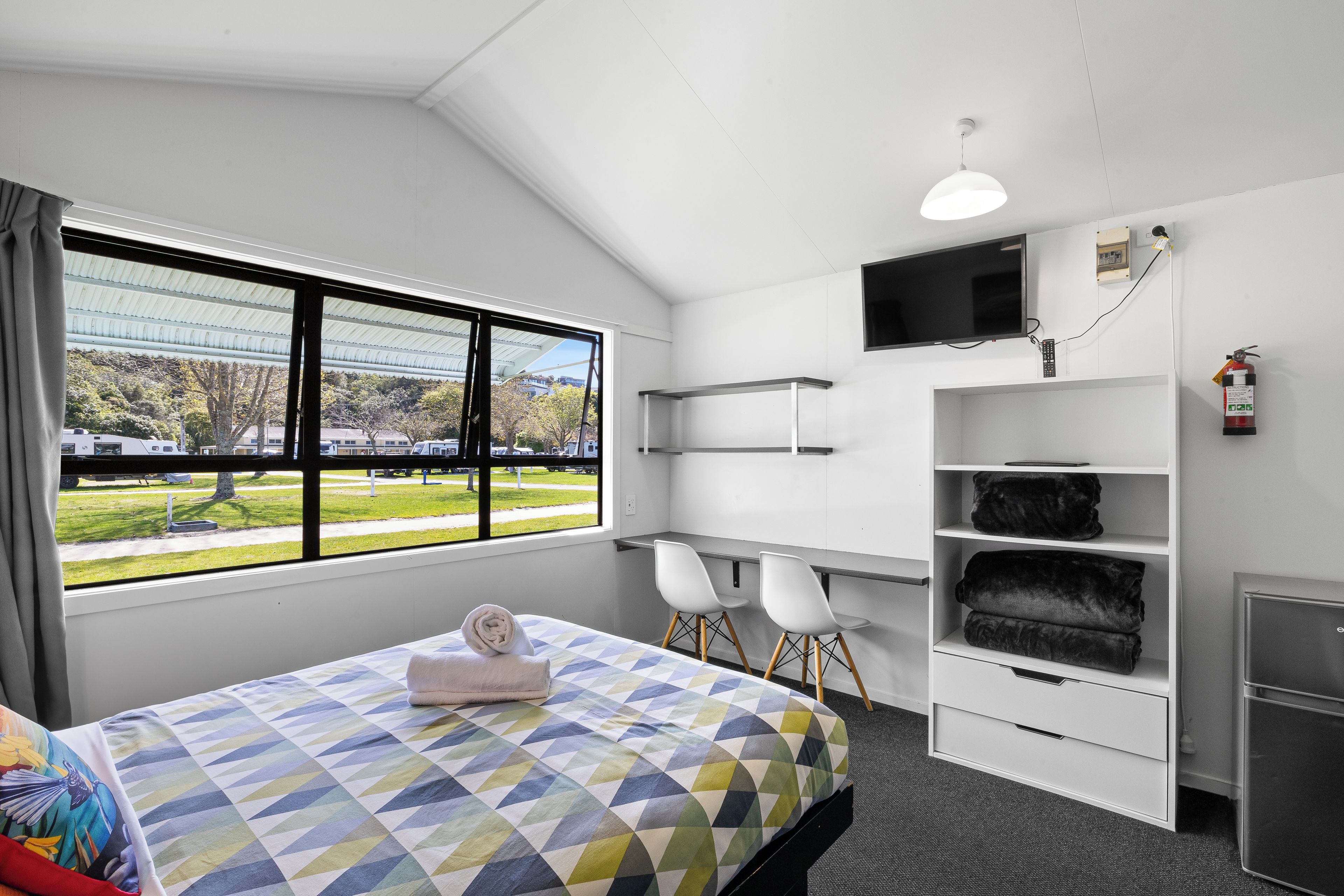 A hotel room with a bed, a TV, a fire extinguisher, and a view of a train.