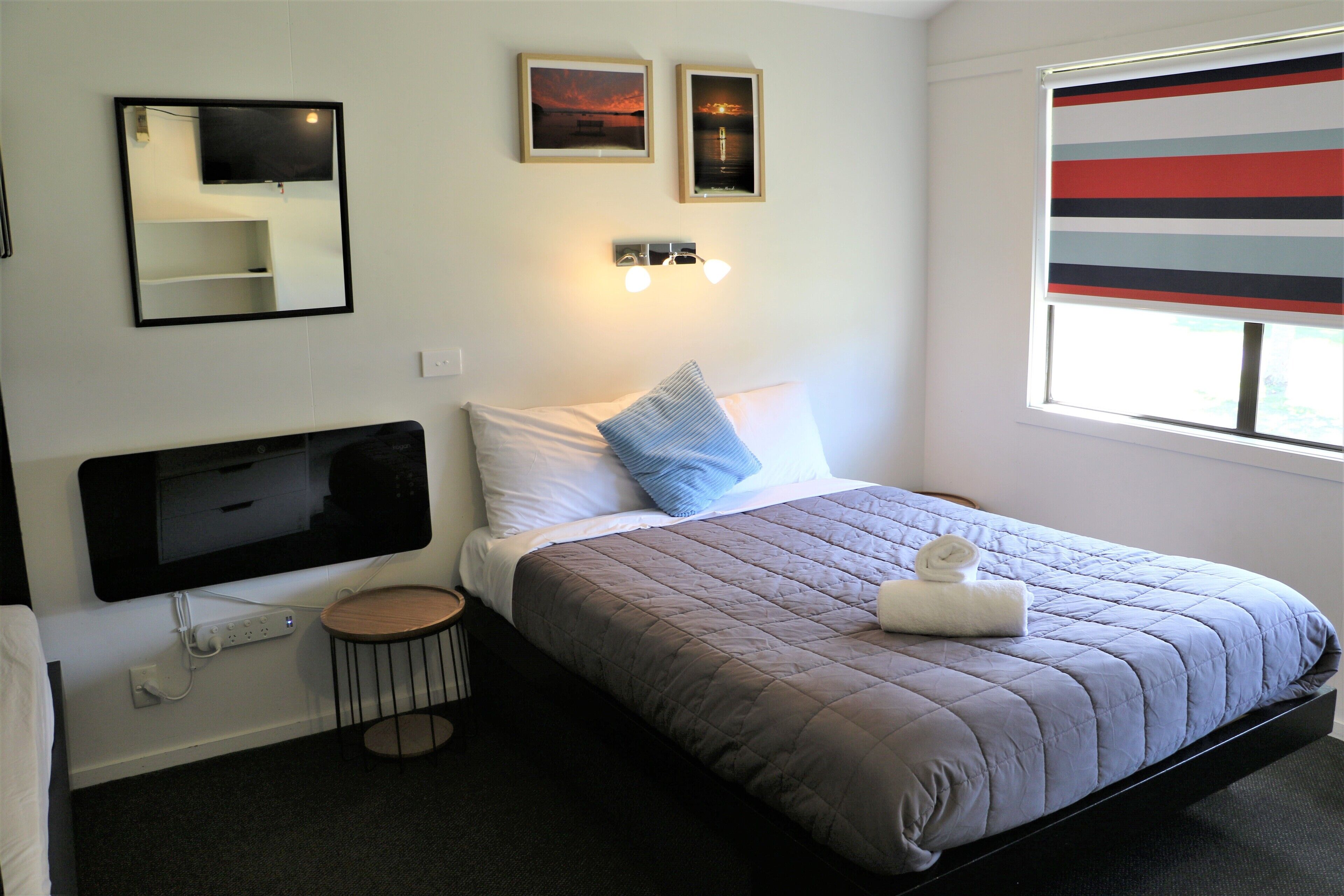 Habitación de hotel con cama, mesita de noche, una mesita redonda, televisor montado en la pared, dos cuadros enmarcados y una ventana con persianas a rayas.
