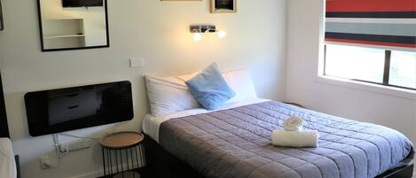 Habitación de hotel con cama, mesita de noche, una mesita redonda, televisor montado en la pared, dos cuadros enmarcados y una ventana con persianas a rayas.