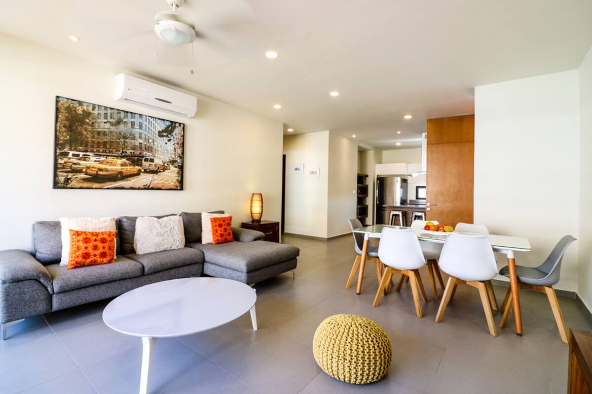 A modern living room with a grey sofa, white coffee table, and a dining area with white chairs and a wooden table.