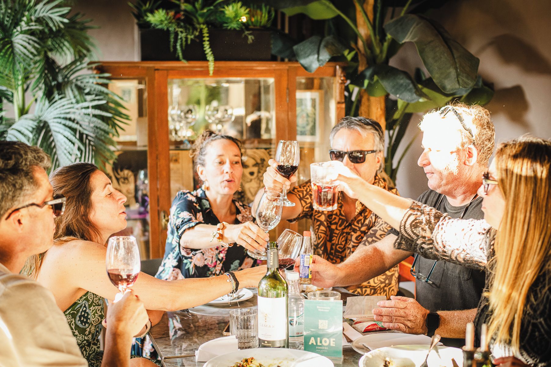 Um grupo de pessoas brindando com taças de vinho à mesa, com uma garrafa e um cardápio.