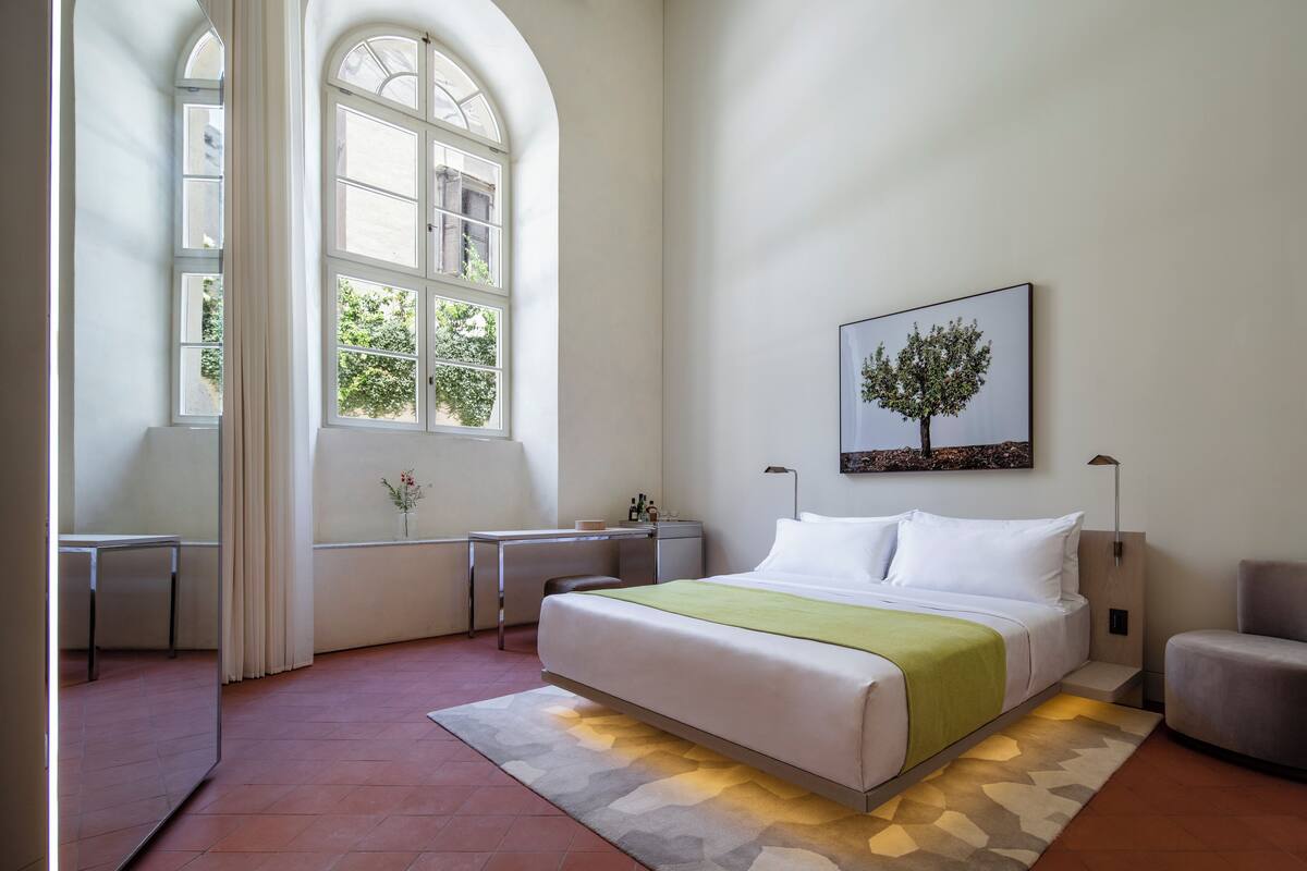 A modern bedroom with a large bed, a desk, and a chair. There is a window with curtains and a painting on the wall.