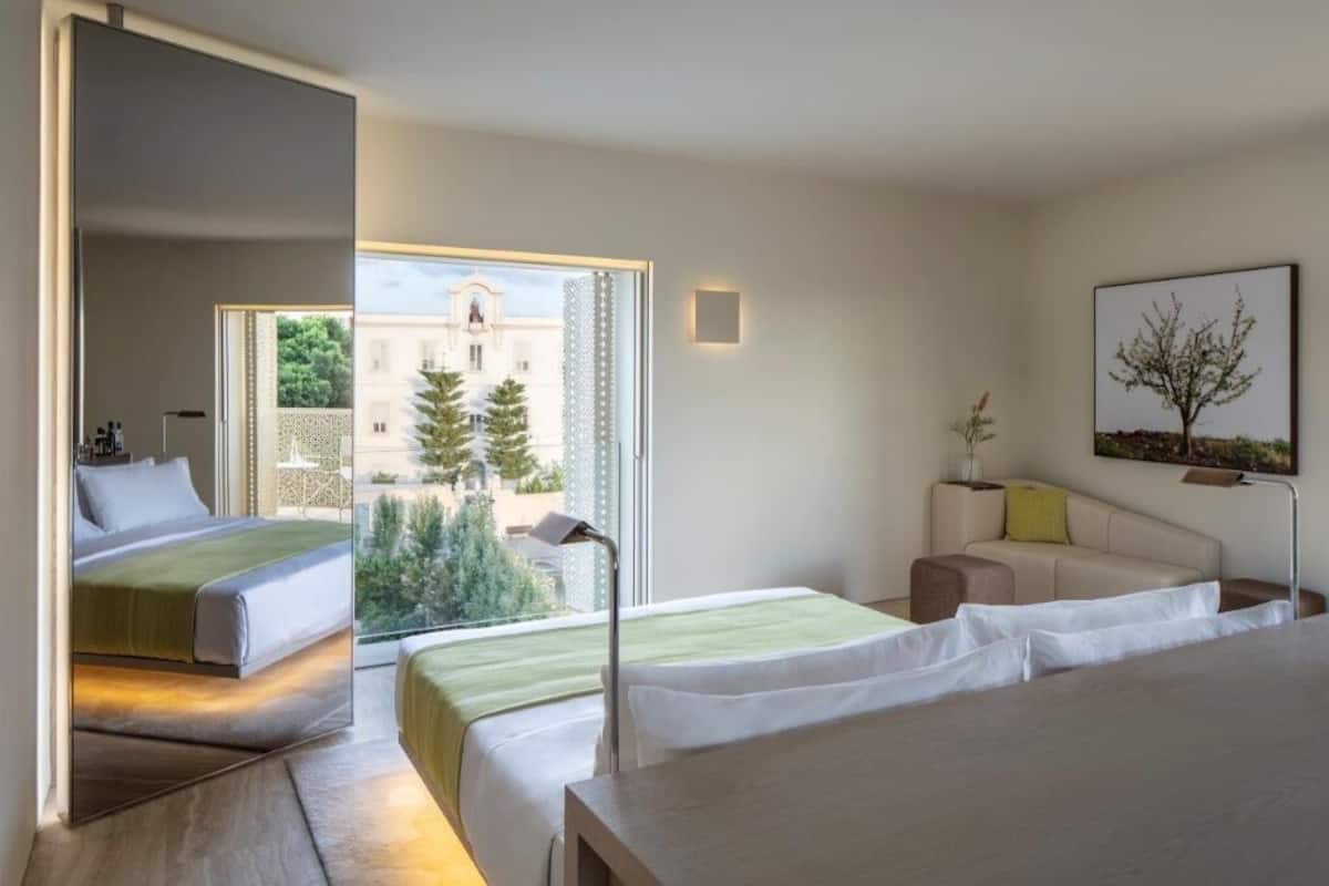 A modern hotel room with two beds, a sofa, a small table, and a balcony with a view of a building and trees.