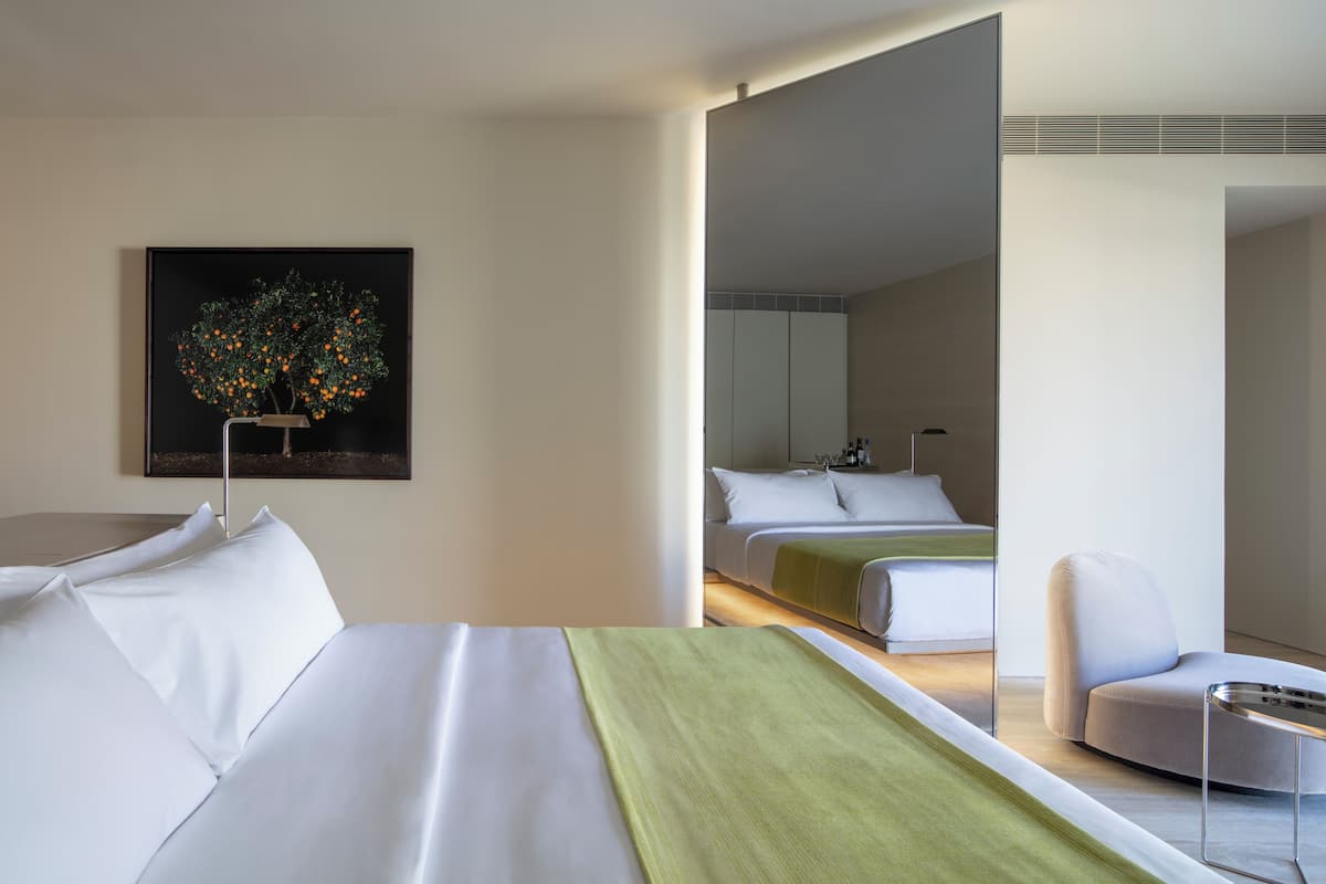 A modern hotel room with a neatly made bed, a green throw blanket, a chair, and a large mirror reflecting the room.