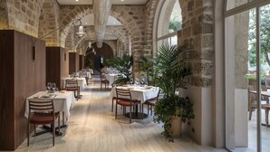 Zona de menjador allargada i estreta amb taules preparades per a diversos comensals, parets de pedra i grans finestrals que deixen passar la llum natural.